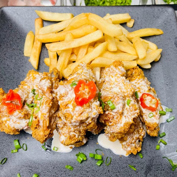 Crispy fried chicken pieces drizzled with white sauce, garnished with red peppers and green onions, served with golden french fries on a dark plate.